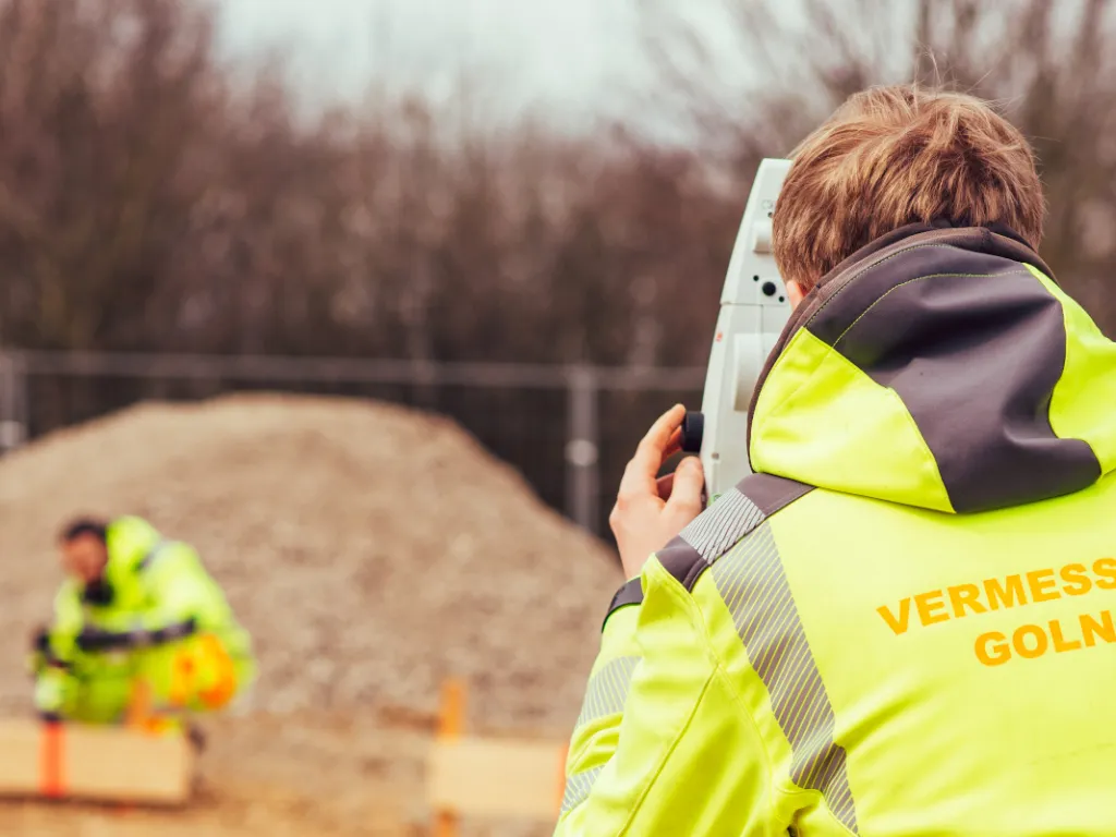 Vermessungsingenieur am Tachymeter auf einer Baustelle, im Hintergrund ein Kollege an der Absteckung