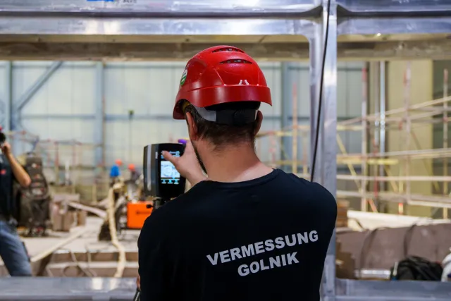 Surveying engineer with measuring instrument in a shipyard hall, a section under construction in the background